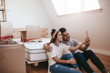 Happy young couple moving in new place and making a selfie. Man and woman sitting on floor with cardboard boxes - JLPSF27238