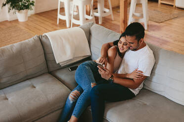 Loving young couple relaxing on sofa and using mobile phone. Young man and woman sitting together on couch and looking at smart phone. - JLPSF27245