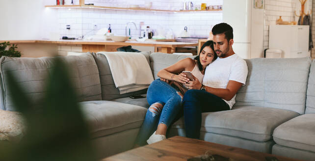 Junges Paar entspannt sich auf der Couch mit Smartphone zu Hause im Wohnzimmer. Mann und Frau sitzen zusammen auf dem Sofa und schauen auf ihr Handy. - JLPSF27246