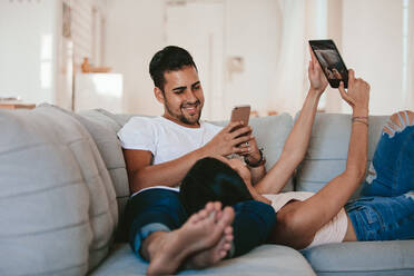 Young man holding mobile phone with woman taking selfie using digital tablet. Young couple relaxing on sofa at home with their digital devices. - JLPSF27249