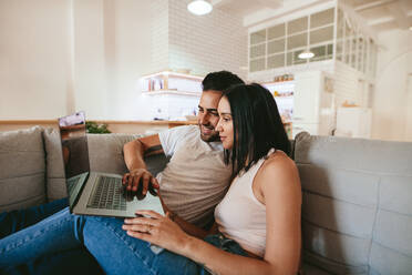 Junges Paar entspannt sich auf der Couch mit Laptop zu Hause im Wohnzimmer. Mann und Frau sitzen zusammen auf dem Sofa und schauen auf den Laptop-Computer. - JLPSF27250