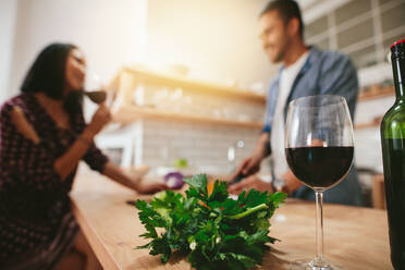 Ein Glas Rotwein auf der Küchentheke mit einem Paar, das im Hintergrund steht. Ein Mann und eine Frau unterhalten sich in der Küche mit einem Glas Wein vor sich. - JLPSF27253