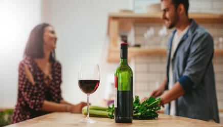 Glas Wein und Weinflasche auf Küchentisch mit Paar Kochen im Hintergrund. Mann und Frau im Gespräch in der Küche mit Wein vor. - JLPSF27254