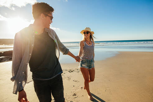 Außenaufnahme eines romantischen jungen Paares, das sich an den Händen hält und am Strand spazieren geht. Junger Mann und Frau gehen an einem Sommertag am Meer spazieren. - JLPSF27266