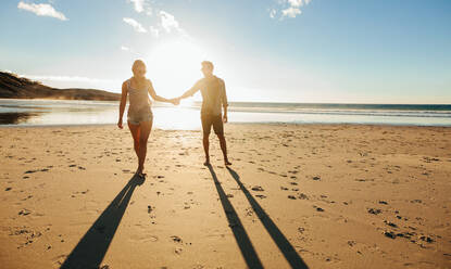 Außenaufnahme eines romantischen jungen Paares, das händchenhaltend am Meer entlang spaziert. Junger Mann und Frau, die bei Sonnenuntergang gemeinsam am Strand spazieren gehen. - JLPSF27270