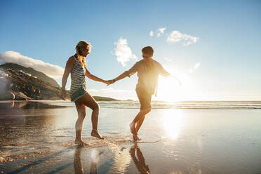 Full length shot of young couple holding hands walking on the beach and having fun. Young man and woman enjoying holidays on the sea shore. - JLPSF27272