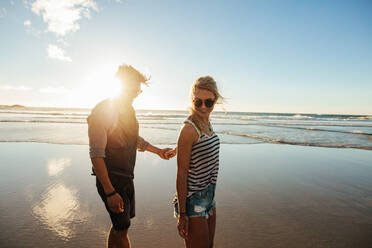 Outdoor shot of romantic young couple walking along the sea shore holding hands. Young man and woman walking on the beach together at sunset. - JLPSF27276