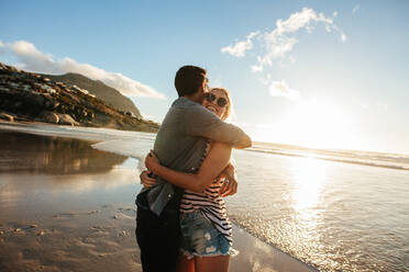 Außenaufnahme eines romantischen jungen Paares, das sich am Strand umarmt. Junger Mann und Frau umarmen sich bei Sonnenuntergang. - JLPSF27277