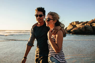 Außenaufnahme eines Paares, das am Strand spazieren geht. Junges glückliches Paar, das am Meer spazieren geht und lächelt. - JLPSF27282