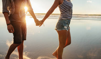 Beschnittene Aufnahme eines jungen Paares, das sich an den Händen hält und am Strand spazieren geht. Liebender Mann und Frau, die am Ufer spazieren gehen. - JLPSF27292
