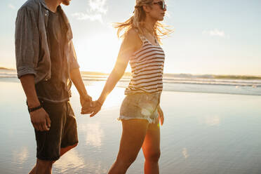 Junger Mann und Frau, die sich an den Händen halten und an einem sonnigen Tag am Strand spazieren gehen. Ein Paar im Sommerurlaub, das am Strand spazieren geht. - JLPSF27293