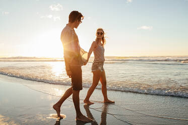 Outdoor shot of romantic young couple walking along the sea shore. Young man and woman walking on the beach together at sunset. - JLPSF27294