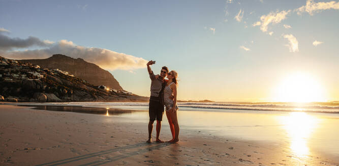 Horizontale Aufnahme eines verliebten jungen Paares, das sich am Strand selbst porträtiert. Mann und Frau am Meer, die ein Selfie mit ihrem Handy machen. - JLPSF27295
