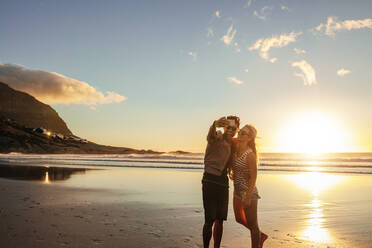 Happy young couple taking self portrait and smiling at the beach. Man and woman enjoying on sea shore taking selfie with mobile phone. - JLPSF27296