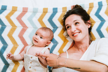 Top view of joyful woman looking at curious relaxed infant with love while resting together on colorful knitted plaid on comfortable bed at home - ADSF39989