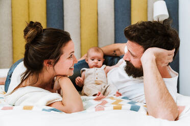 Playful father playing with baby while chilling together with wife and kid on soft bed at home looking at each other - ADSF39995