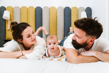 Verspielter Vater, der mit seinem Baby spielt, während er zusammen mit Frau und Kind auf einem weichen Bett zu Hause chillt - ADSF39996