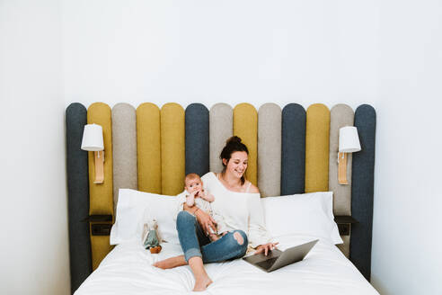 Joyful young woman focusing on screen and interacting with laptop while sitting on soft bed and embracing with love baby against varicolored headboard in light contemporary bedroom - ADSF39997