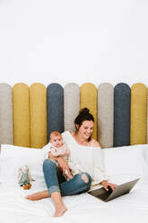 Fröhliche junge Frau, die sich auf den Bildschirm konzentriert und mit dem Laptop interagiert, während sie auf einem weichen Bett sitzt und ihr Baby vor einem bunten Kopfteil in einem hellen, modernen Schlafzimmer umarmt - ADSF39998