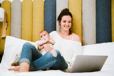 Fröhliche junge Frau, die sich auf den Bildschirm konzentriert und mit dem Laptop interagiert, während sie auf einem weichen Bett sitzt und ihr Baby vor einem bunten Kopfteil in einem hellen, modernen Schlafzimmer umarmt - ADSF40000