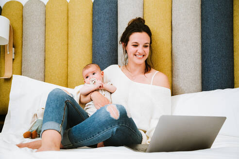 Fröhliche junge Frau, die sich auf den Bildschirm konzentriert und mit dem Laptop interagiert, während sie auf einem weichen Bett sitzt und ihr Baby vor einem bunten Kopfteil in einem hellen, modernen Schlafzimmer umarmt - ADSF40000