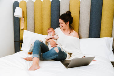 Fröhliche junge Frau, die sich auf den Bildschirm konzentriert und mit dem Laptop interagiert, während sie auf einem weichen Bett sitzt und ihr Baby vor einem bunten Kopfteil in einem hellen, modernen Schlafzimmer umarmt - ADSF40001