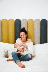 Junge Frau, die ihr Kind liebevoll umarmt und füttert, während sie auf einem weichen Bett vor einem mehrfarbigen Kopfteil in einem hellen modernen Schlafzimmer sitzt - ADSF40002