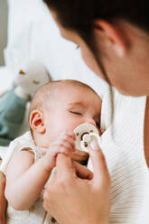 Cropped unrecognizable woman embracing with love and feeding infant while sitting on soft bed against multicolored headboard in light modern bedroom - ADSF40003