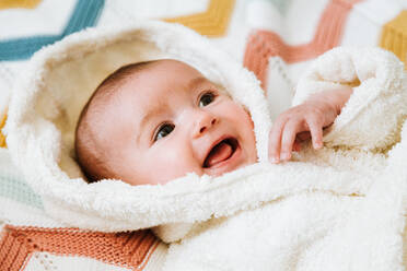 Baby in weichem weißen Bademantel liegt auf bunten gestrickten Plaid auf Bett zu Hause - ADSF40005