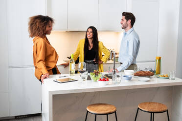 Happy multiracial friends in casual clothes talking and laughing while standing together at kitchen table during home party - ADSF40010