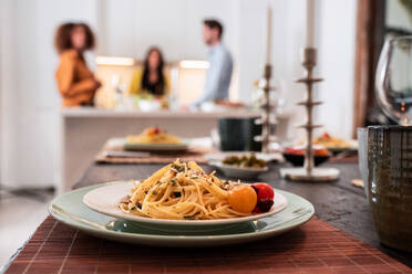 Delicious homemade pasta served on plate on table prepared for home party with group of friends standing together in background - ADSF40011