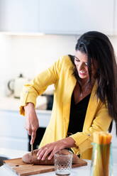 Lächelnde junge Frau in stilvollem Outfit, die am Küchentisch steht und Brot schneidet, während sie Snacks für Freunde während einer Hausparty vorbereitet - ADSF40013