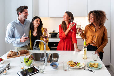 Positive junge gemischtrassige Freunde versammeln sich am Tisch und trinken Wein, während sie die gemeinsame Zeit während einer Hausparty in einer modernen hellen Wohnung genießen - ADSF40015