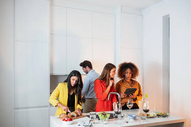 Afroamerikanische Frau, die ein Tablet benutzt, während sie mit Freunden an einem Tisch mit Getränken und Snacks steht und gemeinsam Spaß hat und eine Hausparty in einer modernen hellen Wohnung genießt - ADSF40017