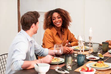 Cheerful romantic multiracial couple flirting while sitting at table and having dinner with friends during home party - ADSF40024