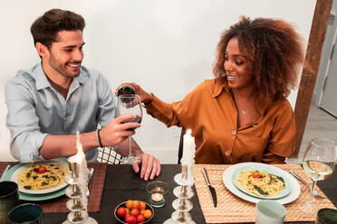 Smiling young African American woman pouring wine while male friend holding glass and together sitting at served table with delicious dishes during dinner at home - ADSF40026