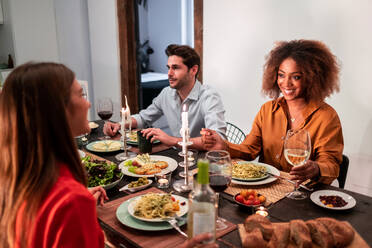 Side view of cheerful multiracial friends sitting at festive table and eating pasta during celebration and party in modern apartment - ADSF40029
