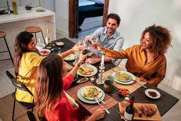 Von oben Seitenansicht von fröhlichen, verschiedenen Freunden, die feiern und mit Weingläsern anstoßen, während sie an einem gedeckten Tisch mit einem köstlichen Abendessen zu Hause sitzen - ADSF40030