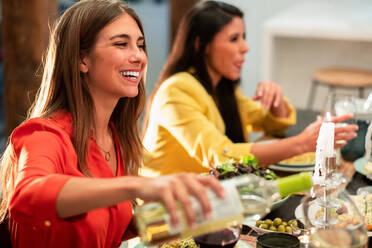 Seitenansicht einer fröhlichen Frau, die Weißwein in einem Glas serviert, mit verschiedenen Freunden, die an einem gedeckten Tisch mit köstlichem Abendessen zu Hause sitzen - ADSF40031