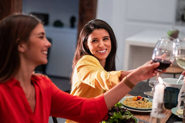 Seitenansicht eines fröhlichen, bunt gemischten Freundeskreises, der feiert und mit Weingläsern anstößt, während er an einem gedeckten Tisch mit einem köstlichen Abendessen zu Hause sitzt - ADSF40033