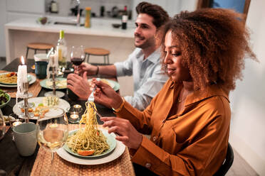 Seitenansicht einer jungen Afroamerikanerin, die Spaghetti isst, während sie mit Freunden beim Abendessen zu Hause am Tisch sitzt - ADSF40037