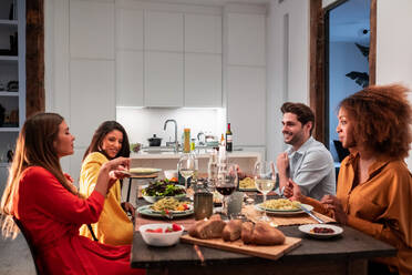 Eine Gruppe junger, unterschiedlicher Freunde sitzt an einem festlichen Tisch und genießt leckeres Essen, während sie das Wochenende zusammen verbringen und zu Hause zu Abend essen - ADSF40038
