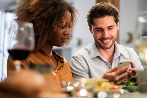 Glückliches junges gemischtrassiges Paar, das während des Abendessens zu Hause auf dem Handy in sozialen Medien surft - ADSF40041