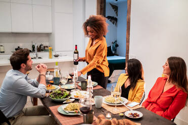 Schwarze afroamerikanische Frau serviert Wein an fröhliche jugendliche Freunde, die lachen und den Zeitvertreib genießen, während sie sich zum Abendessen zu Hause versammeln - ADSF40043