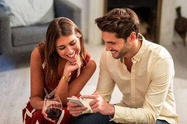 Fröhlicher junger Mann teilt Fotos auf dem Handy mit seiner Freundin, während er sich zu Hause auf dem Sofa ausruht - ADSF40049