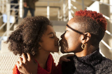 Fröhliches afroamerikanisches Kind küsst seine glückliche Mutter auf der Straße an einem sonnigen Tag - ADSF40109