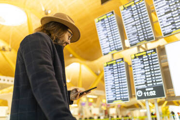 Von unten Seitenansicht der erwachsenen bärtigen männlichen Reisenden in stilvollem Outfit hält Handy und Überprüfung von Informationen über Flug auf Zeitplan Bord im Flughafen - ADSF40118