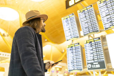 Von unten Seitenansicht der erwachsenen bärtigen männlichen Reisenden in stilvollem Outfit hält Handy und Überprüfung von Informationen über Flug auf Zeitplan Bord im Flughafen - ADSF40119