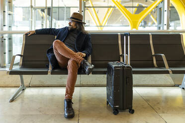 Moderner erwachsener bärtiger Mann in stilvollem Outfit und mit Brille, der auf einem Stuhl neben einem Koffer sitzt und auf einen Flug im Flughafen wartet - ADSF40121
