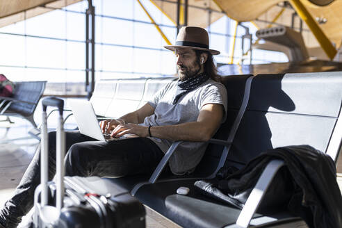 Seitenansicht von fokussierten Mann mit langen Haaren in stilvollem Outfit mit Hut sitzen in einer Bank neben Glaswand arbeiten an einem Laptop während des Wartens auf Flug in Flughafen-Lounge - ADSF40131